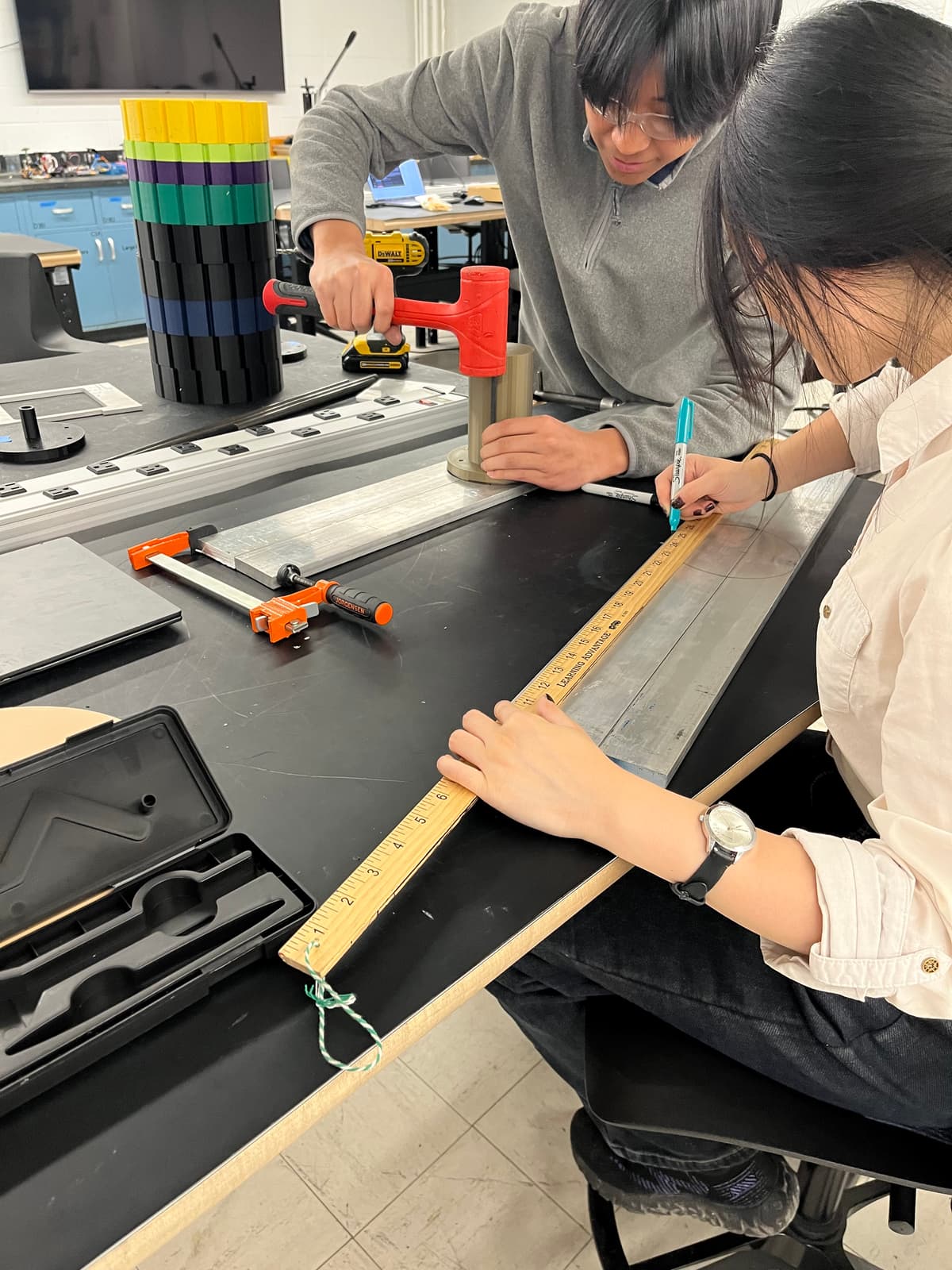 Two team members marking and measuring an aluminium extrusion against a rule on a workbench.