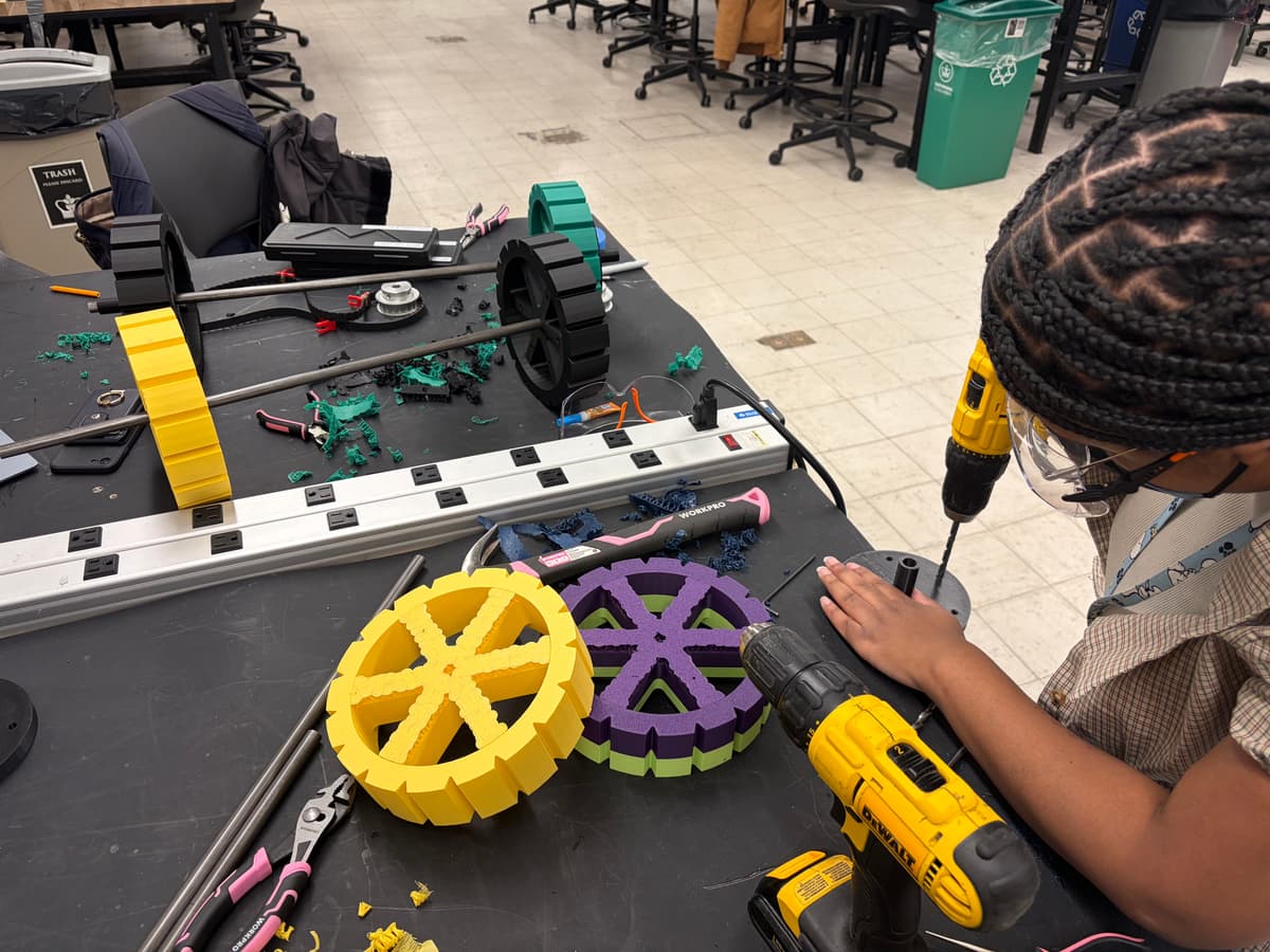 A team member drilling a printed wheel hub at a workbench beside other wheel prototypes.